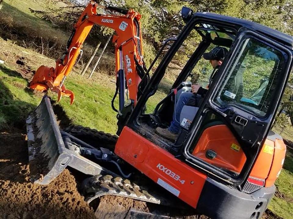 Operator running an orange Kubota KX033-4 mini excavator digging a fence post line in a rural Fraser Valley pasture