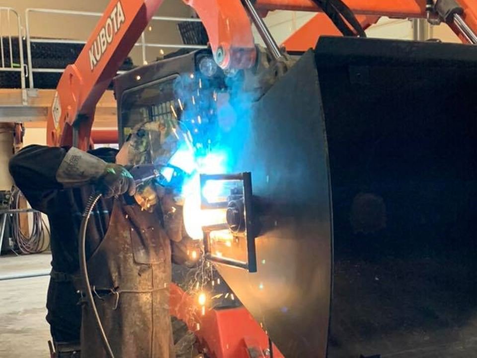 LS Fencing welder MIG welding a custom steel attachment on a Kubota skid steer inside the fabrication shop in Chilliwack